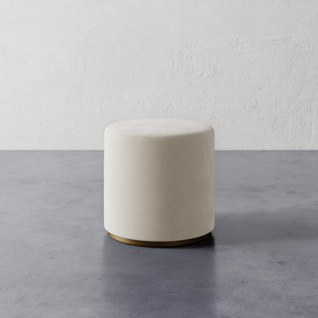 Lemieux et Cie Sophie Round Ottoman in ivory, cylindrical ottoman with brass base, shown on concrete floor against textured plaster walls.