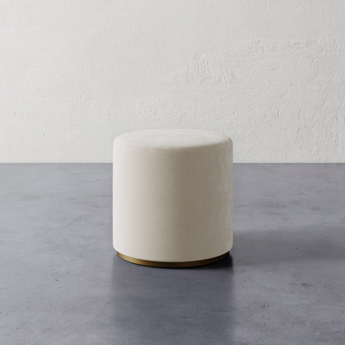 Lemieux et Cie Sophie Round Ottoman in ivory, cylindrical ottoman with brass base, shown on concrete floor against textured plaster walls.