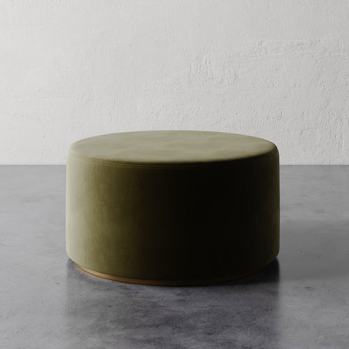 Lemieux et Cie Sophie Round Ottoman in olive green upholstery with brass base trim, low-profile seating on concrete floor against textured plaster wall.