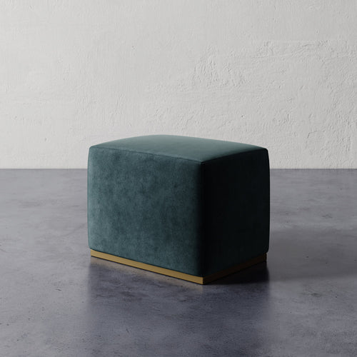 Lemieux et Cie Sophie Rectangular Ottoman in deep teal upholstery with brushed brass base, compact seating piece on polished concrete floor against textured plaster walls.