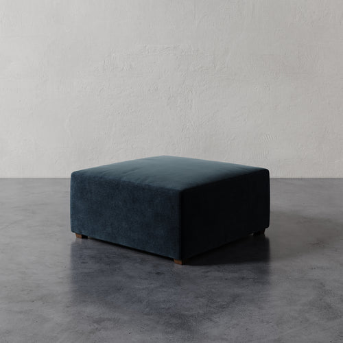 Lemieux et Cie Fabien 36" Square Ottoman in deep teal upholstery, low-profile ottoman with wood legs, shown on concrete floor against textured plaster walls.