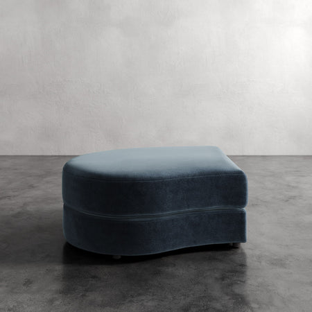 Lemieux et Cie Rulmann Left Ottoman in deep blue, rounded low-profile ottoman on concrete floor against neutral plaster wall.
