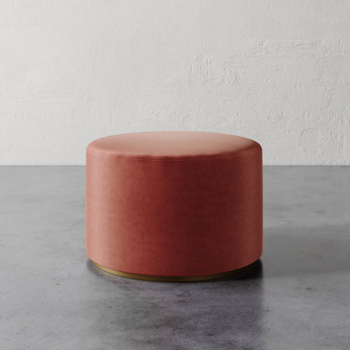 Lemieux et Cie Sophie Round Ottoman in terracotta velvet with brass base, cylindrical ottoman on concrete floor against textured plaster wall.