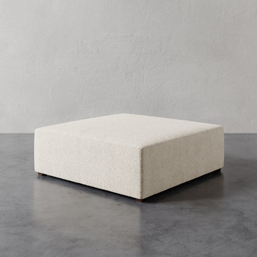 Lemieux et Cie Fabien 48" Square Ottoman in ivory, low-profile square ottoman on polished concrete floor against textured plaster wall.