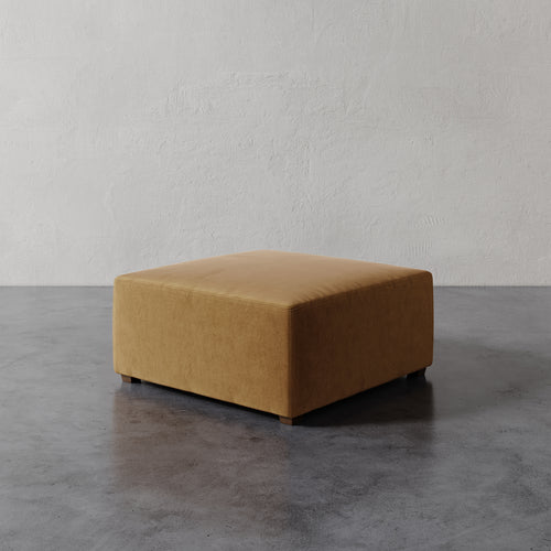 Lemieux et Cie Fabien 36" Square Ottoman in warm camel upholstery, low-profile square ottoman on stone floor against textured plaster wall.