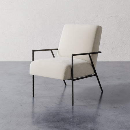 Lemieux et Cie Imane Lounge Chair with ivory upholstery and dark slender frame, lounge chair on polished concrete floor against textured white plaster wall.