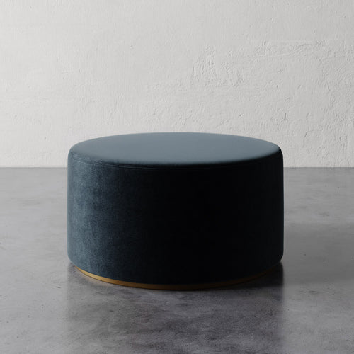 Lemieux et Cie Sophie Round 36" Ottoman in dark teal upholstery with brass base, low-profile round ottoman on concrete floor against textured plaster walls.