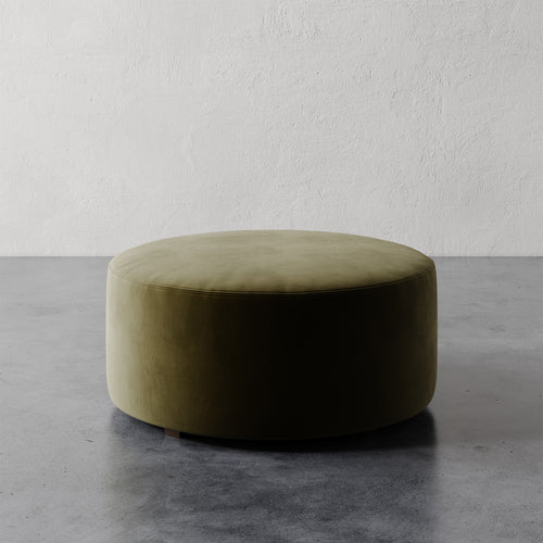 Lemieux et Cie Fabien 36" Round Ottoman in olive green upholstery, low-profile circular ottoman on stone floor against textured plaster walls.