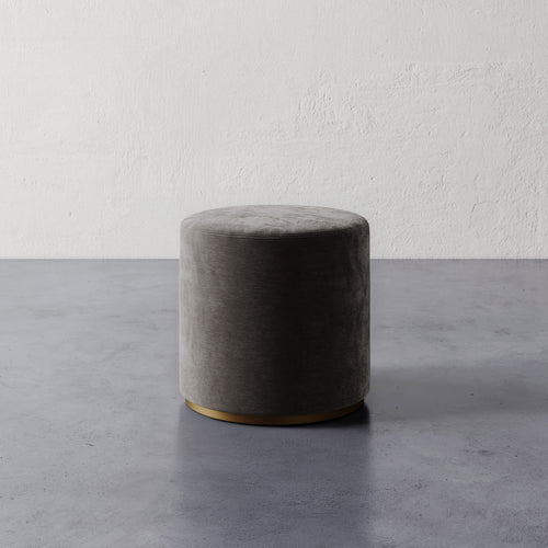 Lemieux et Cie Sophie Round Ottoman in charcoal upholstery with brass base, cylindrical ottoman shown on concrete floor against textured plaster wall.