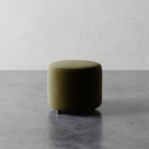 Lemieux et Cie Fabien 22" Round Ottoman in olive green upholstery, low cylindrical ottoman on stone floor against textured plaster walls.