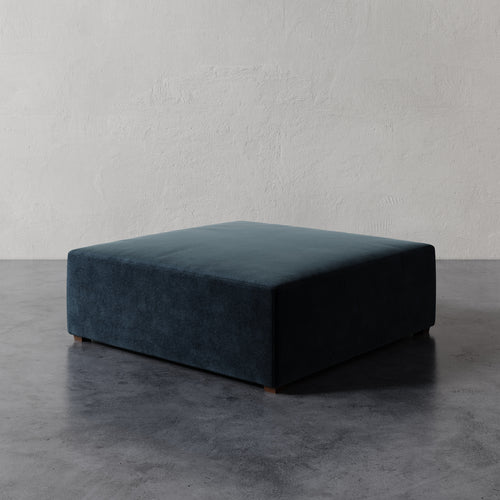 Lemieux et Cie Fabien 48" Square Ottoman in deep teal, low-profile upholstered ottoman on dark concrete floor against textured plaster wall.