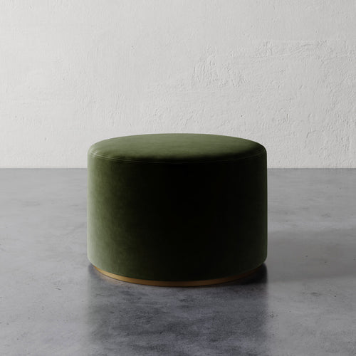 Lemieux et Cie Sophie Round Ottoman in olive green velvet with brass base, low cylindrical ottoman on concrete floor against textured plaster wall.