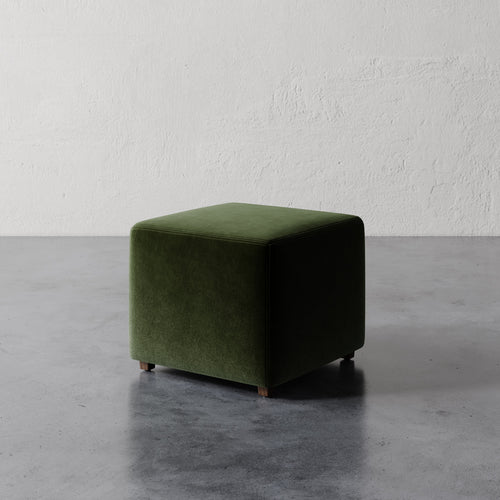 Lemieux et Cie Fabien 22" Square Ottoman in deep green upholstery, handmade accent piece on concrete floor against textured plaster wall.