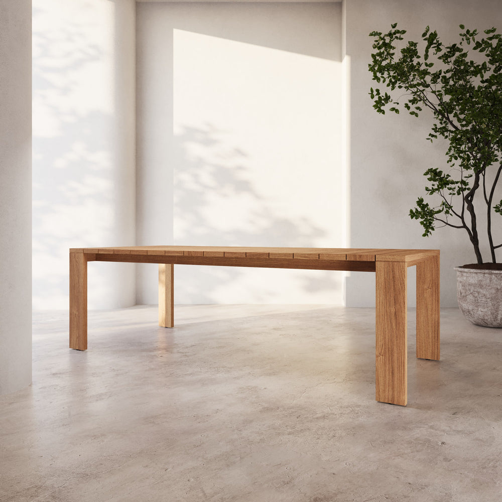 Lemieux et Cie S2 Teak 96" Rectangular Dining Table in solid teak wood with geometric legs, shown on concrete floor in white-walled room with soft daylight and potted tree.