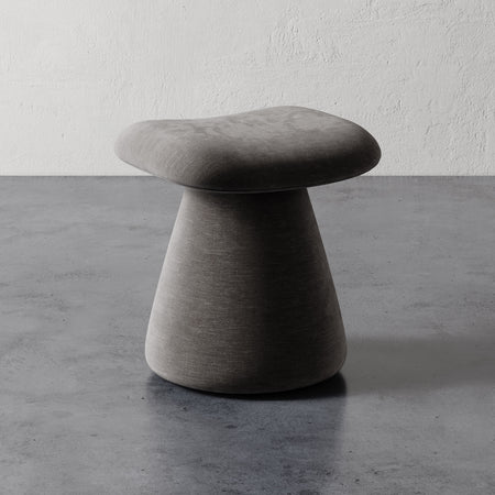 Lemieux et Cie Romy Ottoman, upholstered stool with contoured seat and flared pedestal base, shown on concrete floor against textured plaster walls.