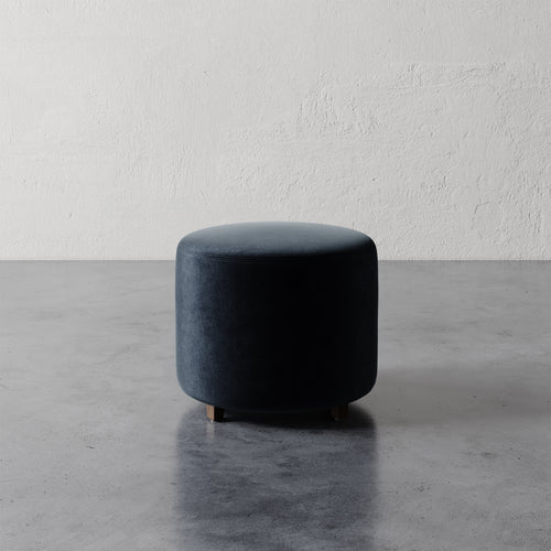 Lemieux et Cie Fabien 22" Round Ottoman in dark upholstery, low-profile cylindrical ottoman on polished concrete floor against textured plaster walls.
