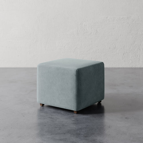 Lemieux et Cie Fabien Square Ottoman in soft sage upholstery, compact cube-form ottoman on concrete floor against textured plaster walls.