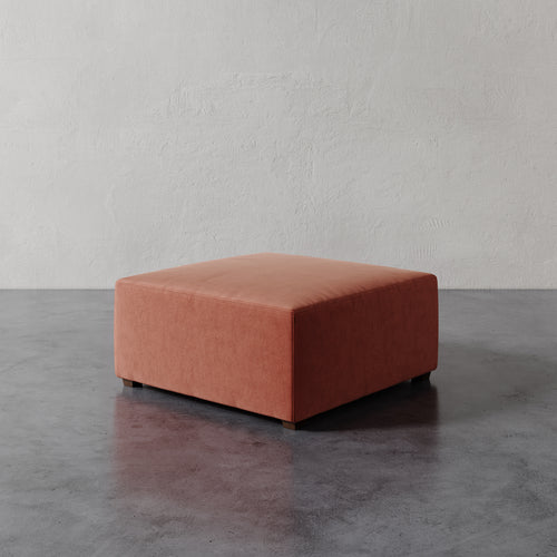 Lemieux et Cie Fabien 36" Square Ottoman in terracotta upholstery, low-profile ottoman on concrete floor against textured plaster wall.