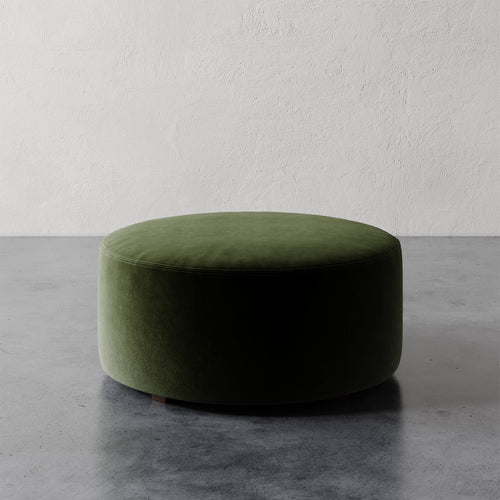 Lemieux et Cie Fabien 36" Round Ottoman in moss green velvet, low-profile circular ottoman on polished concrete floor against textured plaster walls.