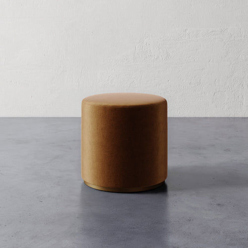 Lemieux et Cie Sophie Round Ottoman in warm caramel upholstery, cylindrical ottoman on concrete floor against textured plaster wall.