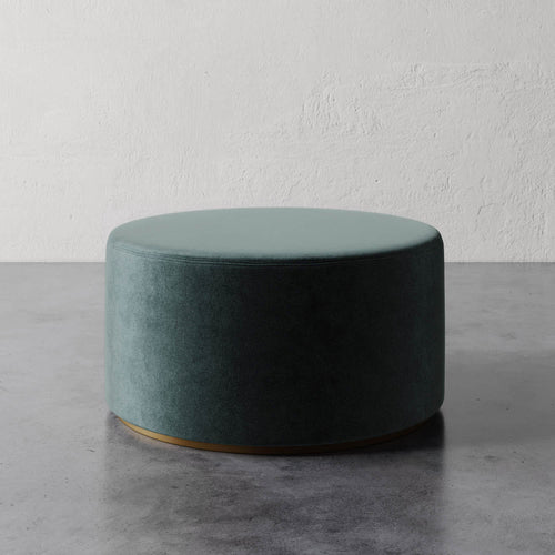 Lemieux et Cie Sophie Round Ottoman in deep teal upholstery with brass base detail, shown on gray concrete floor against textured plaster walls.