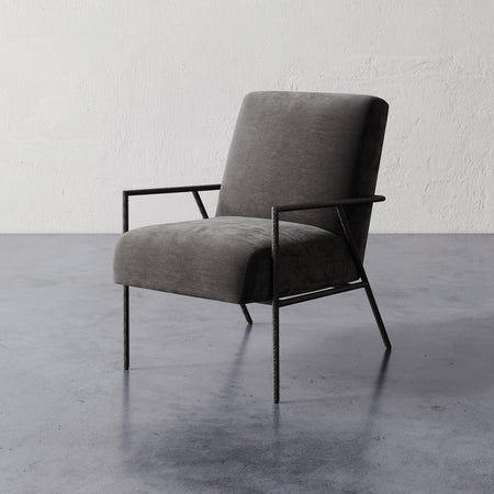 Lemieux et Cie Imane Lounge Chair, dark upholstered chair with slim frame and open arms, on polished concrete floor against textured plaster wall.