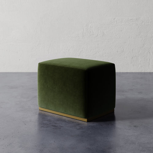 Lemieux et Cie Sophie Rectangular Ottoman in deep green upholstery with brass base, compact ottoman on concrete floor against textured plaster walls.