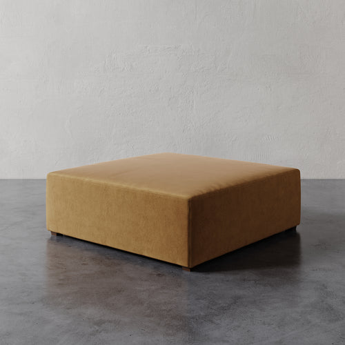 Lemieux et Cie Fabien 48" Square Ottoman in warm caramel, low-profile upholstered ottoman on concrete floor against textured plaster wall.