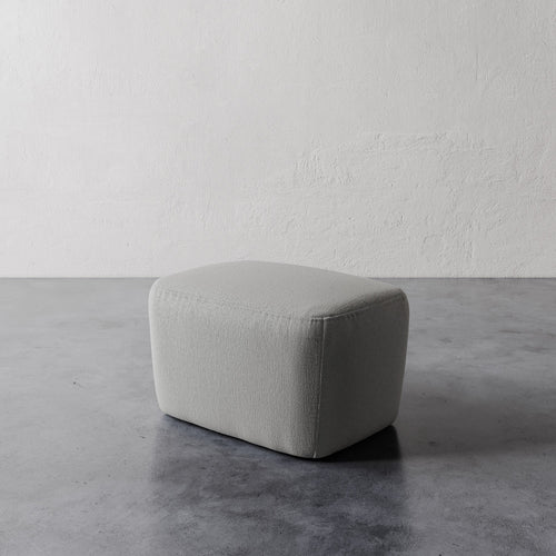 Lemieux et Cie Aurélie Ottoman, rounded low-profile pouf in soft neutral tone, shown on concrete floor against textured plaster walls.