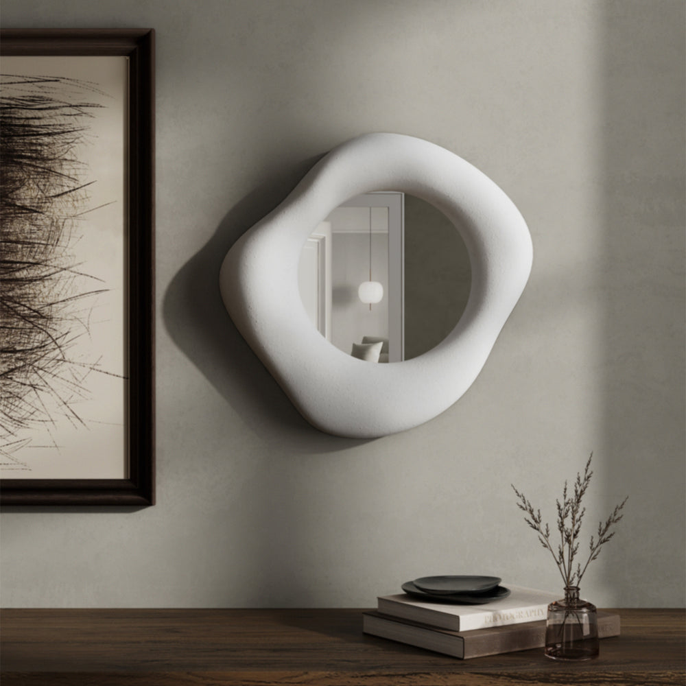 Lemieux et Cie hand-sculpted mirror with asymmetric organic form, sculptural wall-mounted mirror on neutral plaster wall beside framed artwork and wood table with glass vase.