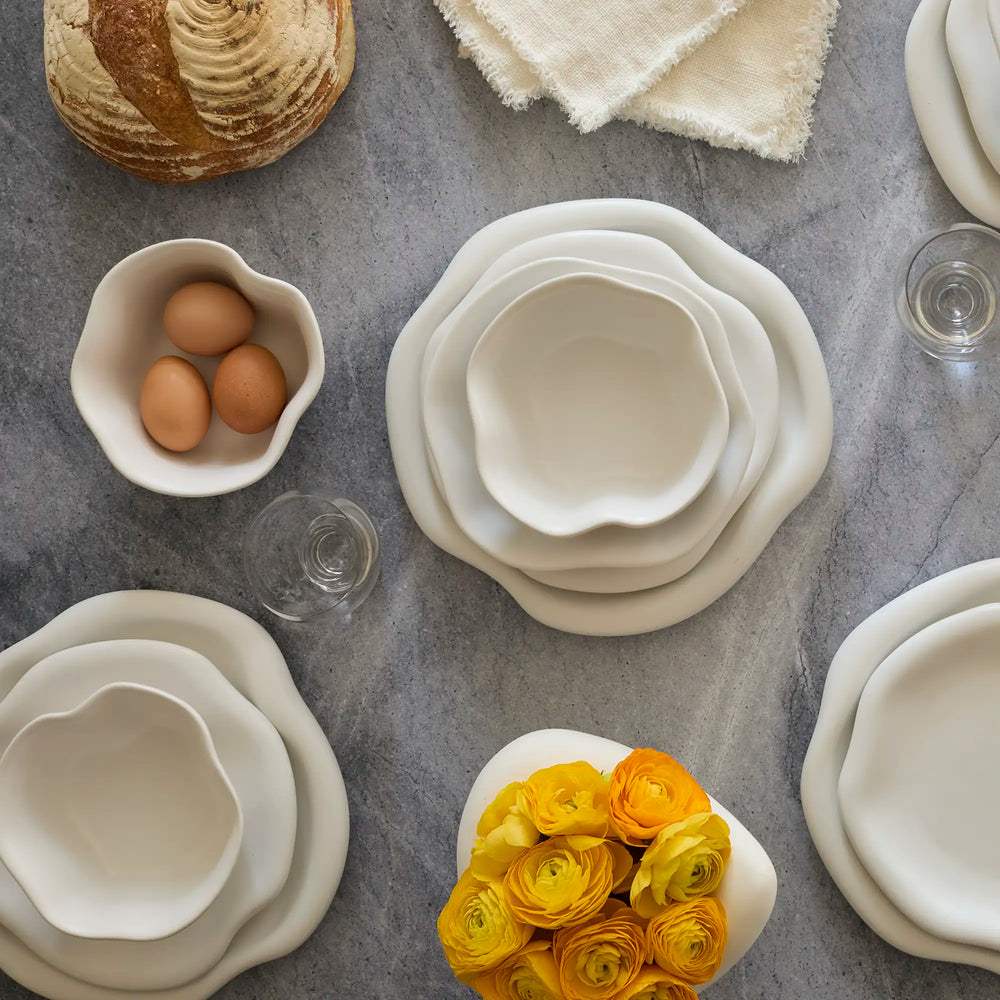 Lemieux et Cie Amorphous Dinnerware Set in soft white matte glaze, stoneware plates with organic asymmetrical edges, arranged on concrete surface with glassware and bread.