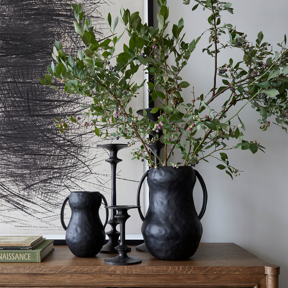 Lemieux et Cie Aquitaine Vase in matte black, organic form with ribbon handles, styled with greenery on wood surface against textured artwork and white walls.