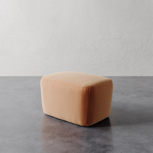 Lemieux et Cie Aurélie Ottoman in warm peach, low rounded ottoman with soft upholstery, set on concrete floor against textured plaster walls.