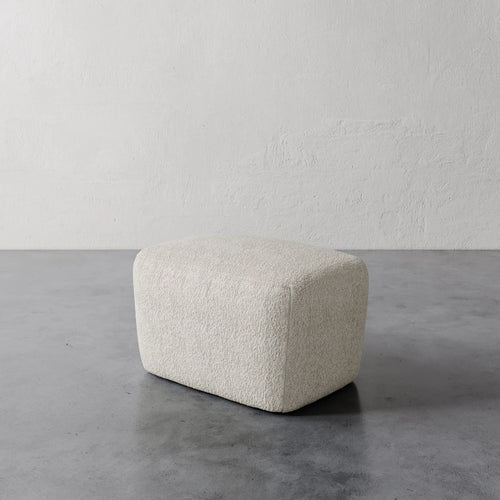 Lemieux et Cie Aurélie Ottoman in cream, rounded low-profile ottoman with soft textured upholstery, shown on concrete floor against plaster wall.