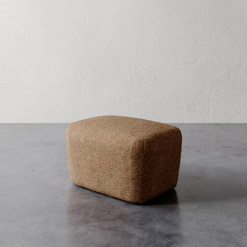 Lemieux et Cie Aurélie Ottoman, low rounded form in warm tan textured upholstery, shown on concrete floor against white plaster walls.