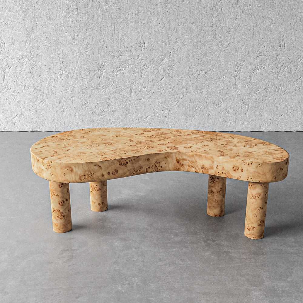 Lemieux et Cie Brenne Burl Coffee Table in Mappa burl veneer, organic kidney-shaped low table with cylindrical legs, on polished concrete floor against textured plaster wall.