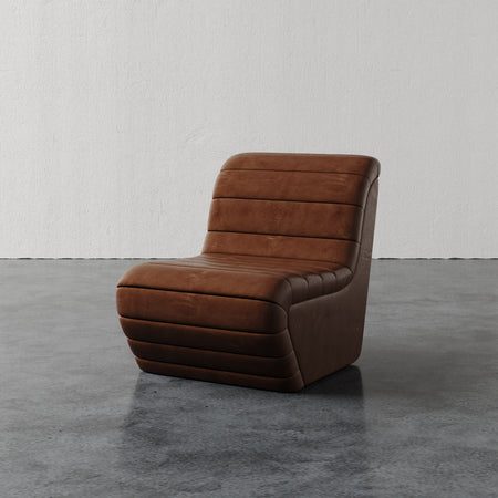 Lemieux et Cie Brunoy Leather Chair in warm brown with channeled upholstery, sculptural slipper chair on concrete floor against plaster wall.