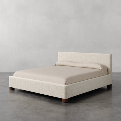 Lemieux et Cie Gérard Bed in ivory bouclé, low-profile platform bed with wrapped headboard and block feet, shown on polished concrete floor against pale plaster walls.