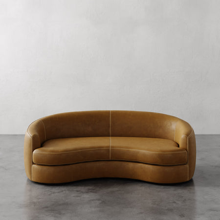 Lemieux et Cie Rulmann Leather Sofa in warm caramel, curved organic sofa with sculptural form, shown on concrete floor against white plaster wall.