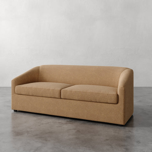 Lemieux et Cie Arvelle Queen Sleeper in warm camel, curved-arm sofa with low-profile base, shown on polished concrete floor against plaster walls.