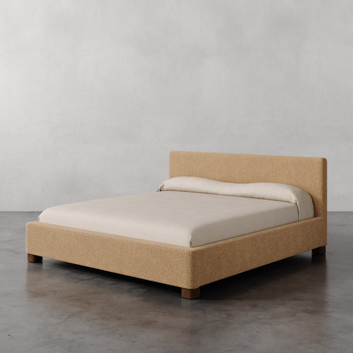 Lemieux et Cie Gérard Bed in warm bouclé upholstery with ivory bedding, low-profile platform bed on dark concrete floor against pale plaster wall.