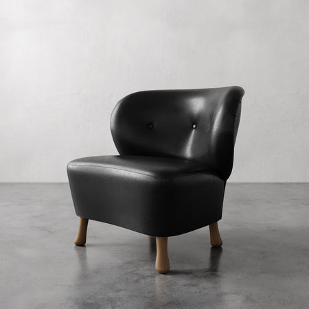 Lemieux et Cie Tourenne Chair in dark leather with button tufting on rounded back, chair on tapered legs, stone floor and neutral walls.