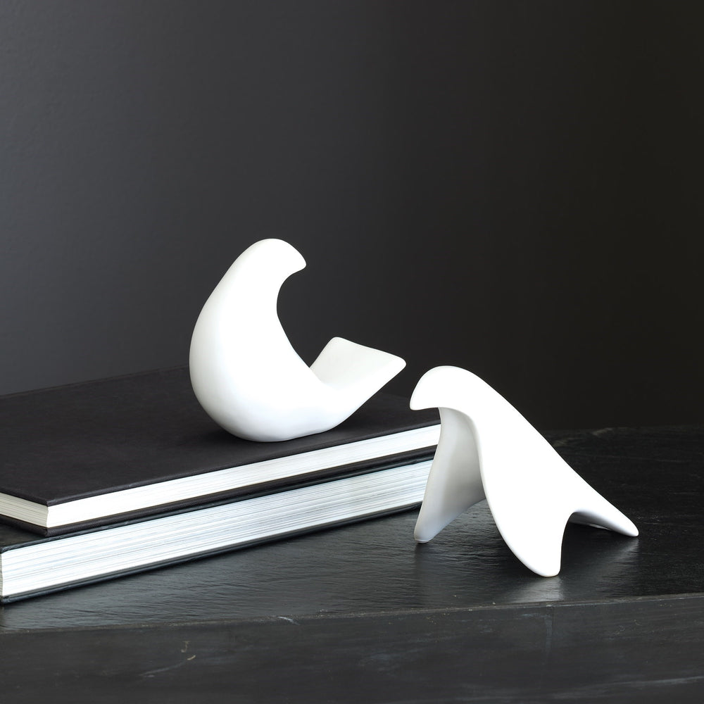 Lemieux et Cie bird objects in white, sculptural forms resting on stacked books atop a dark wood surface against charcoal walls.