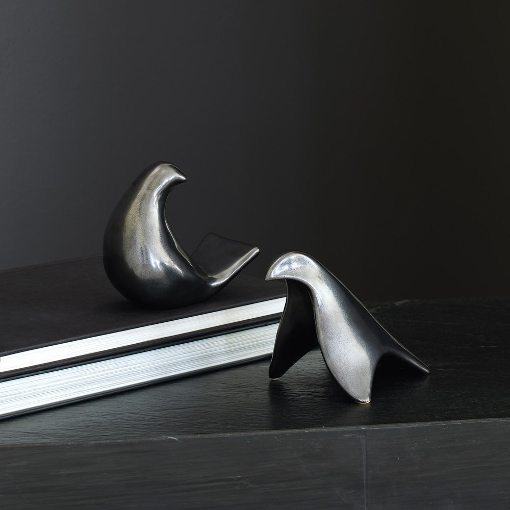 Lemieux et Cie sculptural bird objects in polished dark finish, shown resting on a book atop a dark wood surface against charcoal walls.