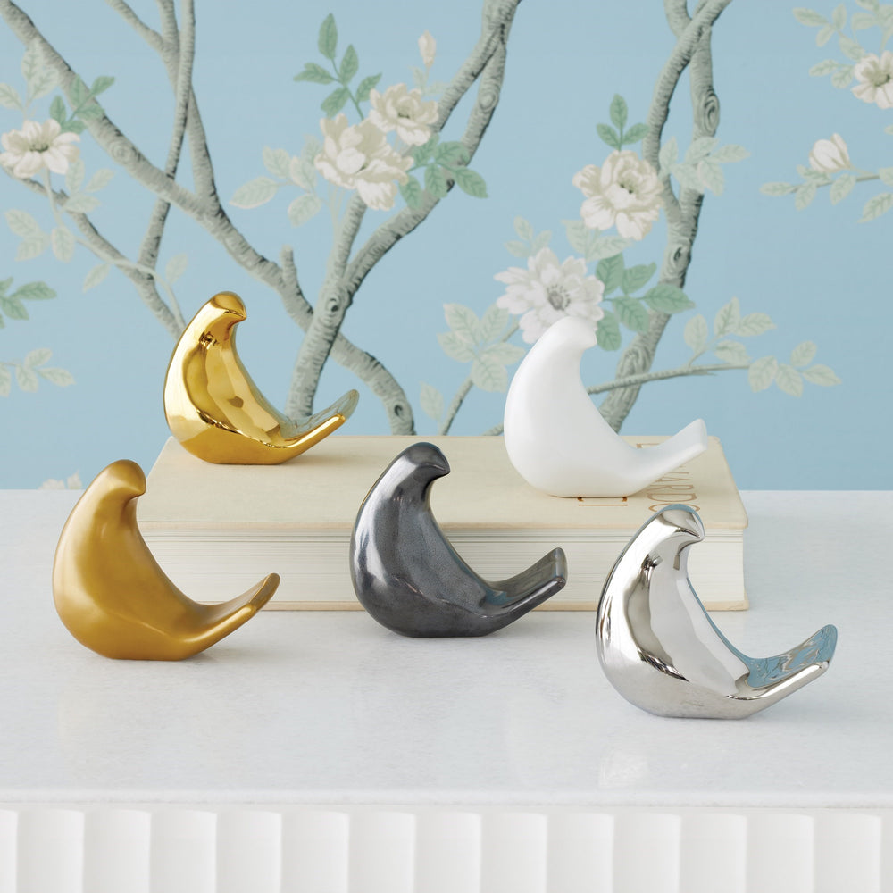 Lemieux et Cie bird objects in polished gold, matte white, charcoal and silver finishes, decorative sculptures arranged on stacked books against hand-painted floral wallpaper.