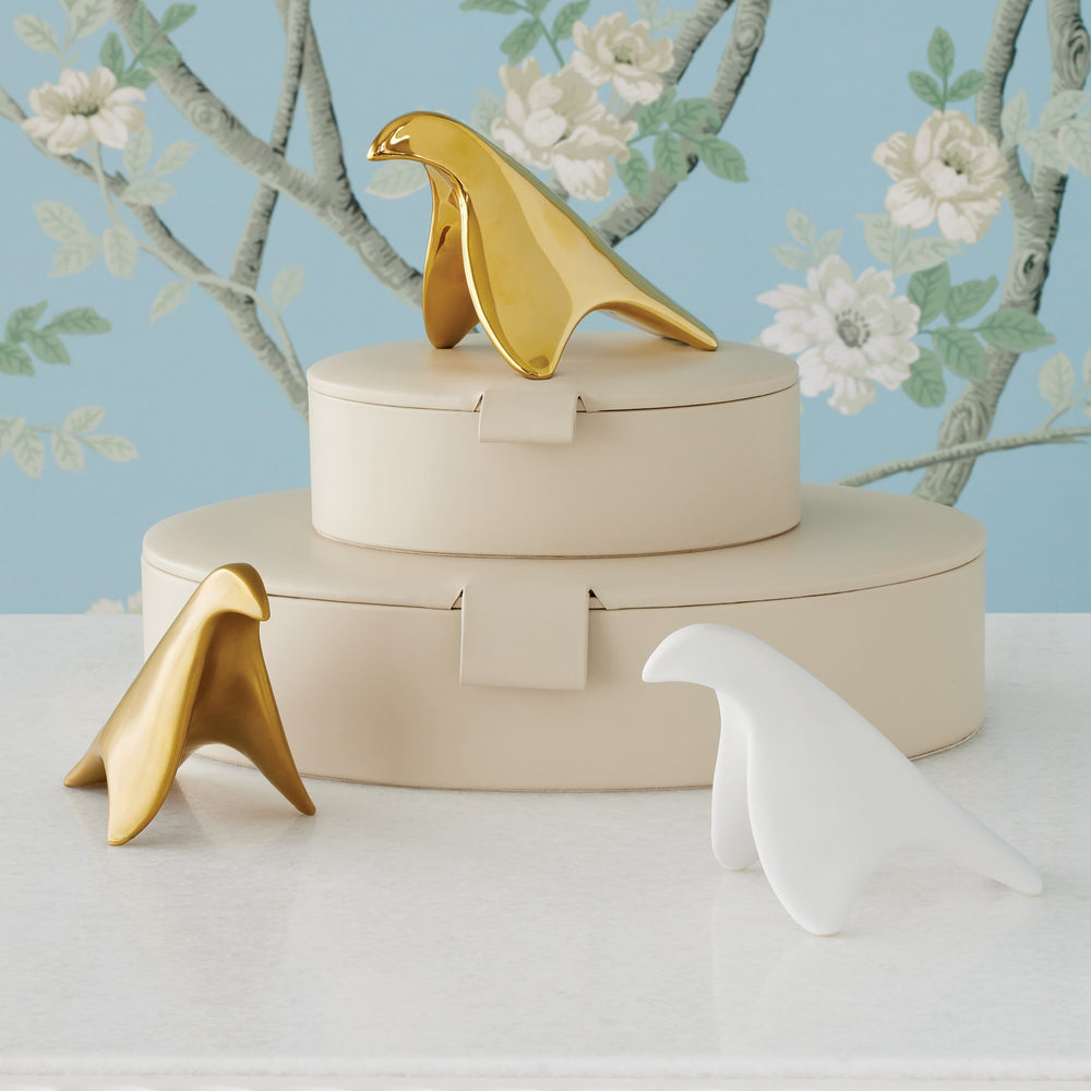 Lemieux et Cie bird objects in polished gold and matte white finishes, sculptural forms displayed on ivory oval boxes against floral wallpaper backdrop.