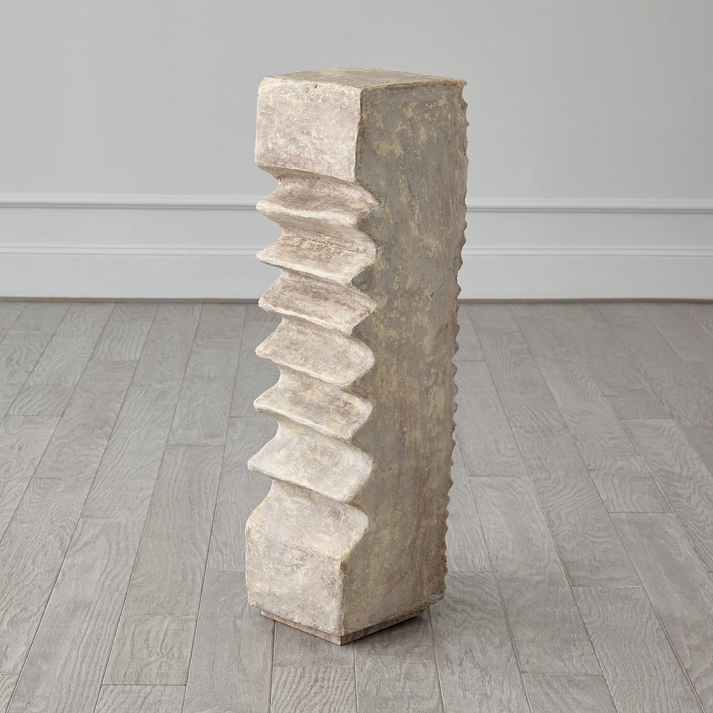 Lemieux et Cie Corentin Candle Holder in dark forged metal, sculptural stepped pedestal with textured finish, shown on light wood floors with neutral walls.