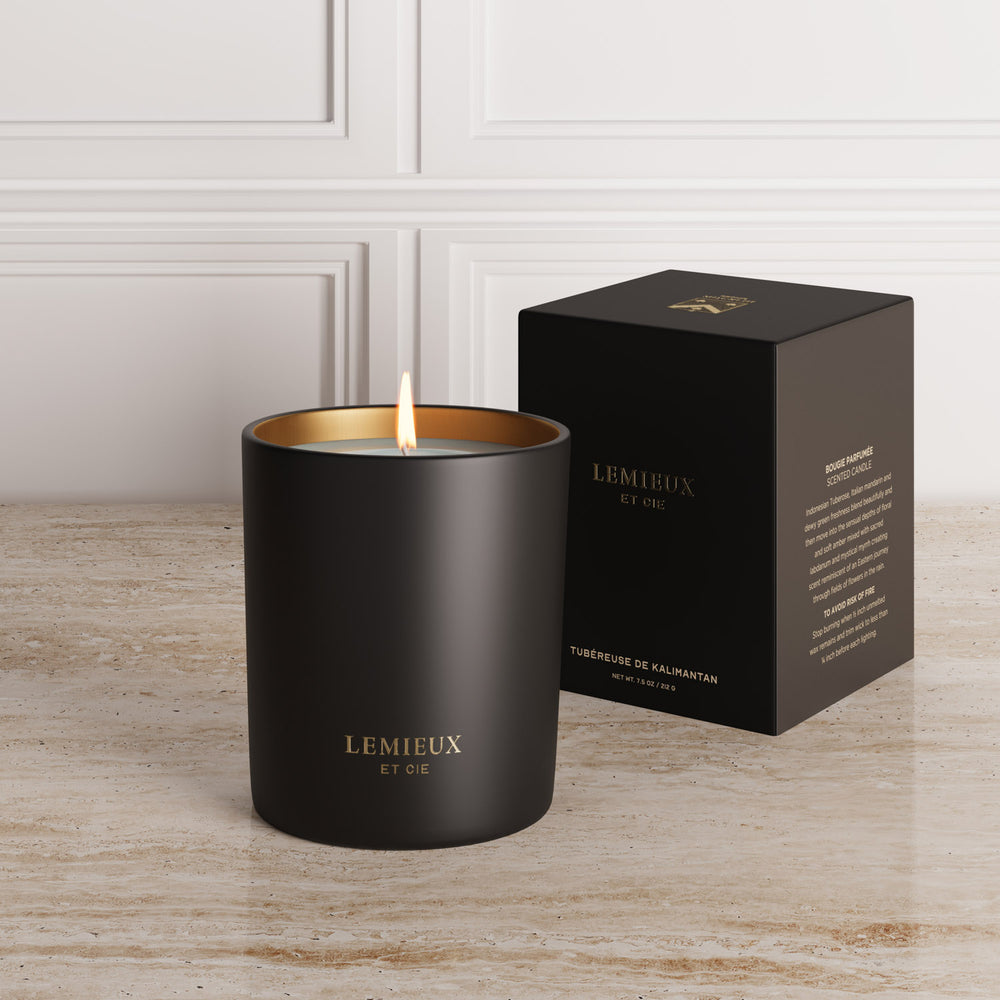 Lemieux et Cie Tubéreuse de Kalimantan scented candle with lit flame, matte black glass vessel beside packaging on stone surface, white paneled walls.