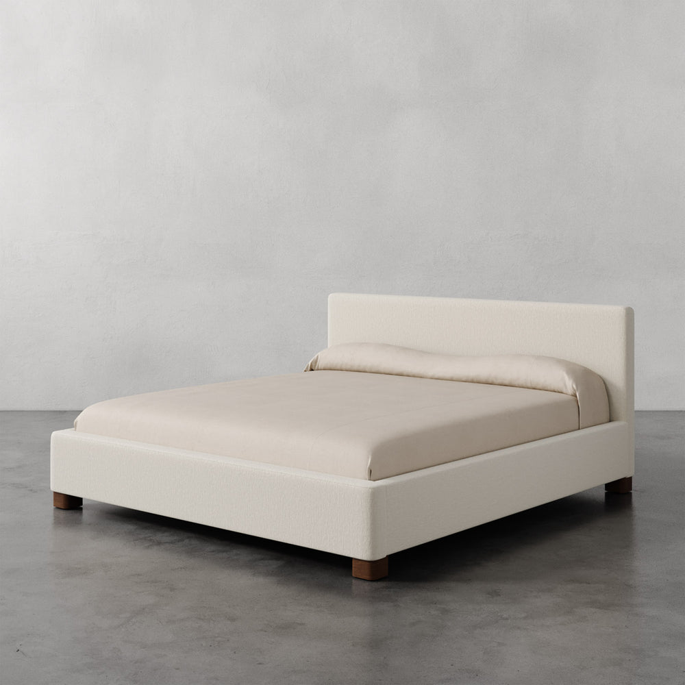 Lemieux et Cie Gérard Bed in ivory upholstery, low-profile platform bed with wrapped headboard and block feet, shown on concrete flooring against neutral walls.