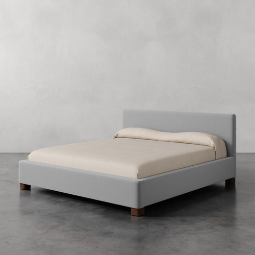 Lemieux et Cie Gérard Bed in pale gray upholstery with rounded block feet, low-profile platform bed dressed in ivory linens, on polished concrete floor.